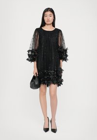 Zwarte jurk met doorschijnende, opgeblazen mouwen; versierd met kant en bloemen appliqués langs de zoom. G gedragen met stijlvolle zwarte hakken en een kleine handtas.