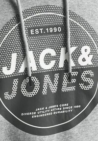 Felpa grigia con cappuccio, caratterizzata da una grafica circolare con il testo "JACK & JONES" in bianco e un motivo esagonale. I lacci del cappuccio sono grigi con puntali metallici.