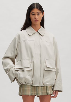 Woman with long dark hair wearing a loose, light beige jacket with large front pockets and a green plaid skirt against a plain background.