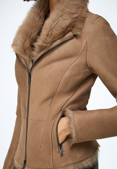 Veste en suédine marron avec un col en fausse fourrure, une fermeture éclair à l'avant, des poches latérales et des coutures détaillées le long des coutures. Texture douce.