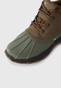 Zapato marrón y verde impermeable con una puntera de goma moldeada y texturizada, y diseño de cordones. Presenta la marca John Deere y una suela duradera.