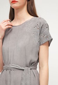 Harmaa mekko, jossa on lyhyet hihat, kukkakirjailuja ja leikkauksia hartioissa. Mukana vastaava vyö.