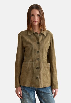 Donna che indossa una giacca di camoscio verde oliva con bottoni e grandi tasche frontali, abbinata a jeans a fantasia floreale blu.