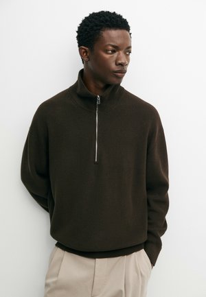 Homme portant un pull à fermeture éclair partielle marron foncé et un pantalon beige, debout devant un fond blanc uni, regardant sur le côté.