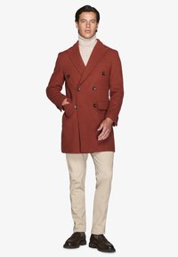 Rustrød dobbeltradet frakke med struktureret stof, der har mørke knapper, sidelommer og spidse lapeller. Sammen med beige bukser.