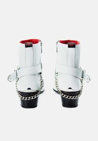 White leather ankle boots with a red interior, featuring a strap and silver chain detail, and a black heel. Zipper on the side for closure.