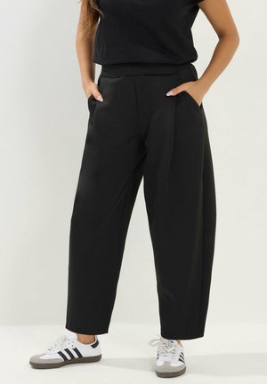 Personne portant un pantalon noir ample et des baskets blanches avec des rayures noires, les mains dans les poches, debout devant un fond clair uni.