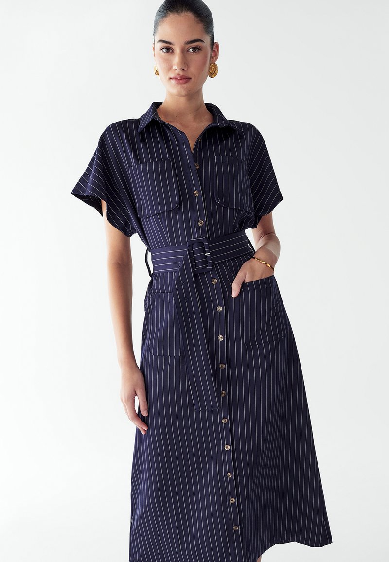 Tumesinine triibuline t-särk-kleit lühikeste varrukatega, nööppluus, kahe rinnaesise taskuga, vöödiga talje ja põlvest allapoole ulatuva pikkusega.