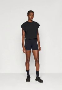 Black cropped t-shirt with wide sleeves, paired with navy shorts. Model wears black shoes and socks, standing on a plain background.
