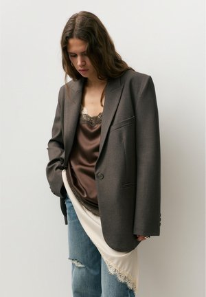 Donna con lunghi capelli castani che indossa un blazer grigio oversize, una canottiera di raso marrone con pizzo, jeans blu strappati e un dettaglio di gonna in pizzo bianco.