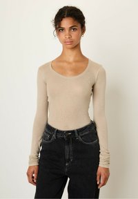 Beige long-sleeve top with a scoop neckline made of textured fabric, paired with high-waisted black jeans featuring contrast stitching and pockets.