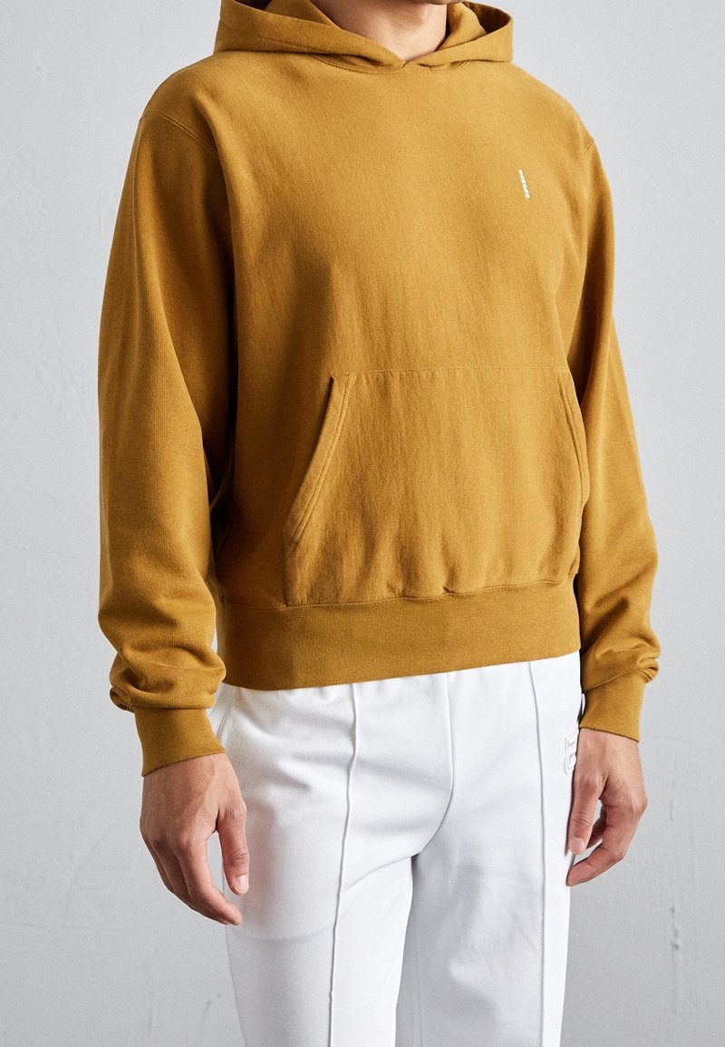 Felpa con cappuccio color giallo senape con tasca frontale, maniche lunghe e cappuccio con coulisse. Tessuto morbido e vestibilità rilassata, indossata sopra pantaloni bianchi.