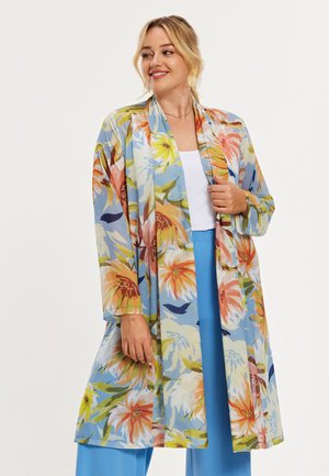 Vrouw draagt een lange, losse kimono-achtige jas met bloemenprint over een witte top en felblauwe broek, glimlacht en kijkt opzij.