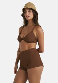 Braunes geripptes Bikini-Set mit triangularer Oberseite und hochtaillierten Shorts; Model trägt einen gewebten beigen Bucket-Hut; glatte Textur.
