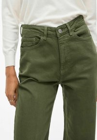 Pantalones de algodón verde oliva con bolsillos delanteros y traseros, cierre con botón de metal y diseño de pierna recta. Tela texturizada visible.