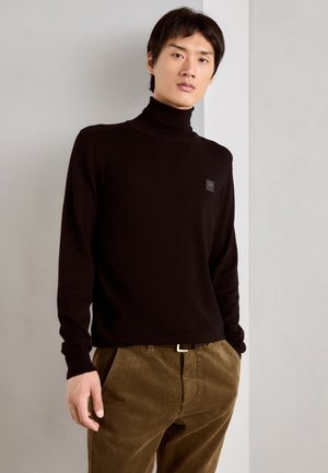 Suéter negro de cuello alto fabricado en un tejido suave, con mangas largas y un pequeño parche de logo. Combinado con pantalones de pana verde olivo.