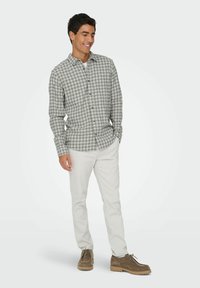 Jeune homme souriant, portant une chemise à carreaux grise, un pantalon blanc et des chaussures en cuir marron à lacets, se tenant les mains dans les poches.