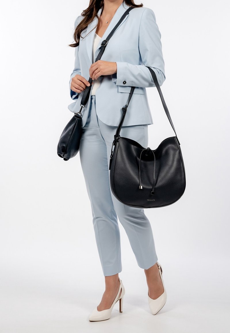 Femme en costume bleu clair portant des talons blancs, tenant deux sacs à bandoulière en cuir noir, debout sur un fond blanc.