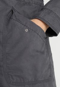 Šedá kapsa na bundě s výrazným patentním knoflíkem, texturovanou tkaninou a viditelným šitím, do které je částečně zasunutá ruka.