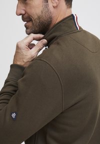 Rjava zip-up jakna z visokim ovratnikom, ki ima rdeče, bele in modre akcentne barve. Mehka tekstura in prilega se telesu s logotipom na rokavu.