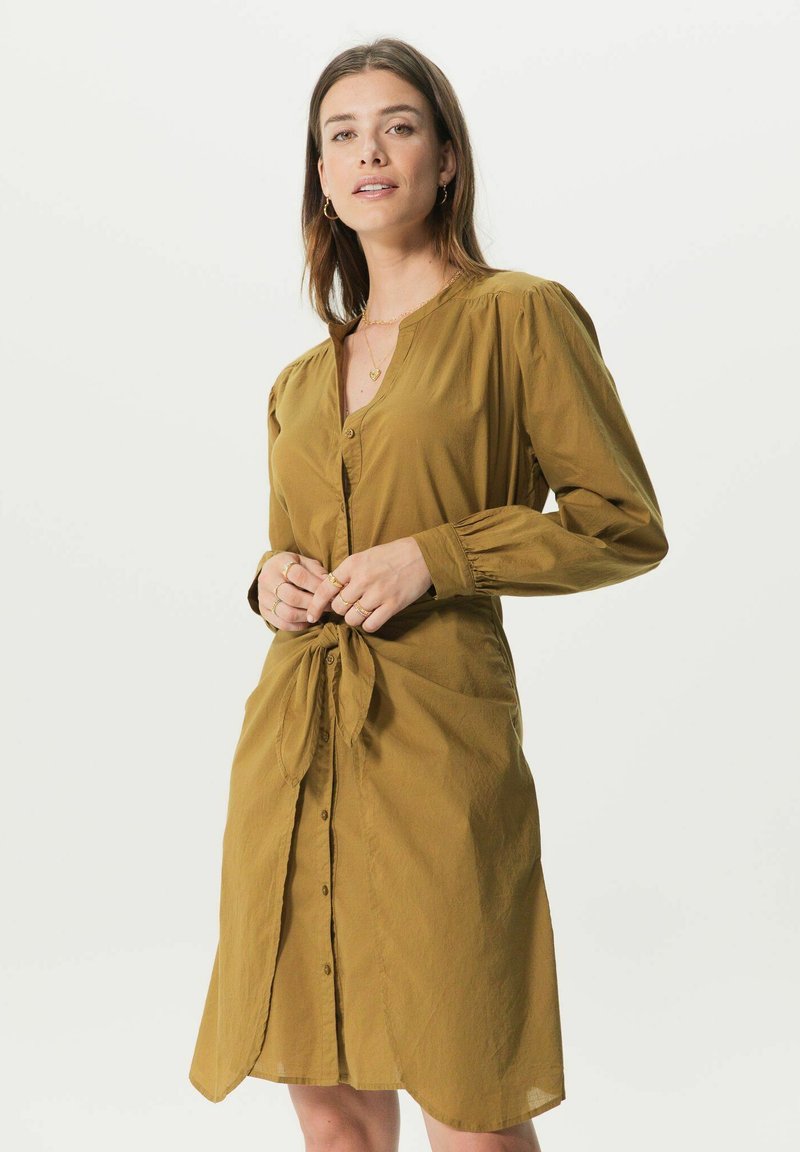 Robe chemise de couleur moutarde avec un devant à boutons, des manches longues et un détail de taille nouée. Le tissu semble léger avec une texture douce.