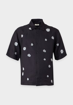 Un homme se tient debout, les mains dans les poches, portant une chemise noire à manches courtes à boutons avec des imprimés floraux blancs, un pantalon ample foncé et des chaussures noires.