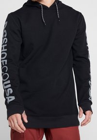 Felpa nera con cappuccio in tessuto morbido, dotata di tasca frontale, cappuccio con coulisse e scritte bianche lungo le maniche. Design semplice e casual.
