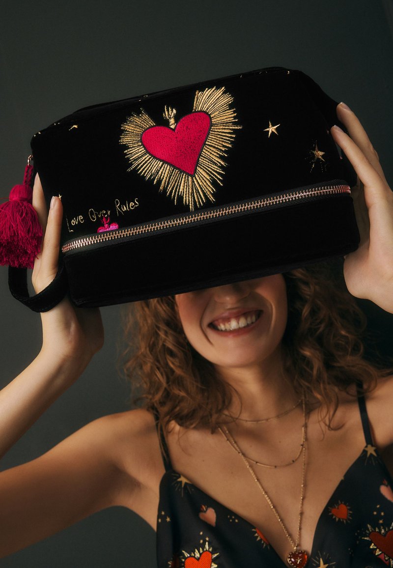 Schwarze Kosmetiktasche aus Samt mit goldener und pinker Herzstickerei, silbernem Reißverschluss und pinkem Quastenakzent. Der Text lautet "Love Over Rules."
