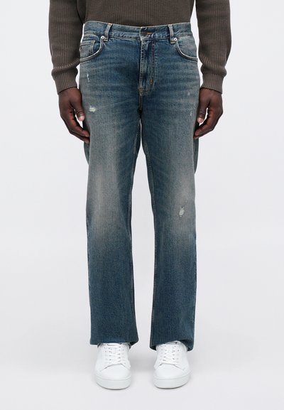 Μπλε τζιν παντελόνι με ίσια γραμμή, με ελαφρές φθορές και κλασικό σχέδιο πέντε τσέπες. Συνδυασμένο με λευκά αθλητικά παπούτσια.