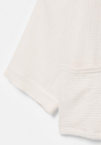 Gros plan sur un tissu blanc texturé montrant les détails de la manche longue et de la poche inférieure d’un vêtement.