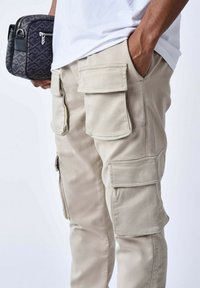 Pantaloni cargo beige con molte tasche a pattina, realizzati in tessuto misto cotone, accanto a una borsa a tracolla nera con motivo.