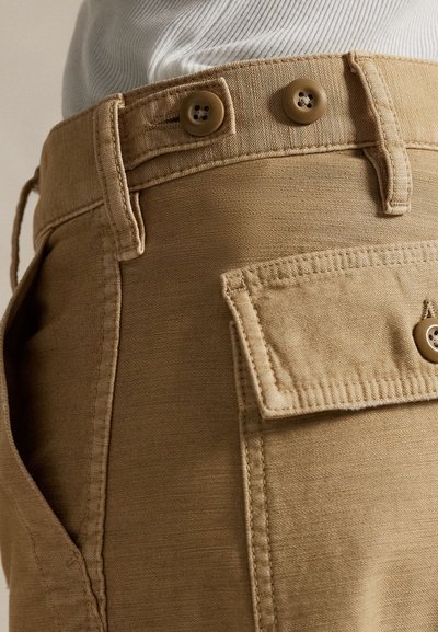 Close-up of tan pants with buttoned waistband and flap pocket, worn with a white ribbed top.