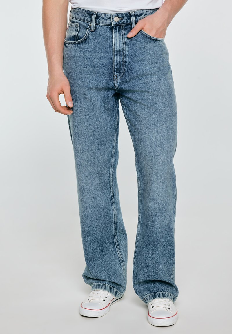 Mężczyzna noszący jasnoniebieskie spodnie jeansowe o prostych nogawkach i białe trampki, z prawą ręką w kieszeni, a lewą luźno opuszczoną wzdłuż boku.