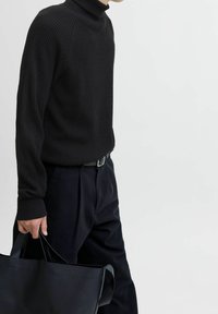 Pull col roulé noir à côtes associé à un pantalon foncé. Le mannequin tient un grand cabas en cuir noir. Design simple et texturé.