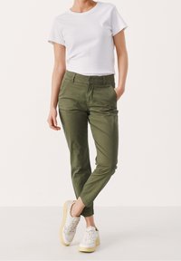 Pantalons en coton vert avec une coupe slim, poches avant et texture lisse, associés à une chemise blanche à manches courtes et des baskets blanches.