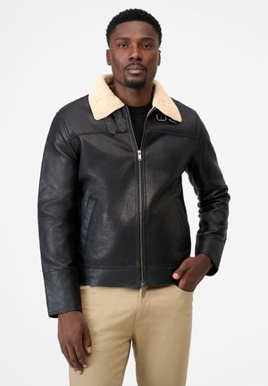 Mann trägt schwarze Lederjacke mit cremefarbenem Lammfellkragen und beige Hose, steht mit einer Hand in der Tasche.