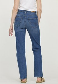 Jean en denim bleu à coupe droite, avec des poches arrière ornées de détails de couture. Porté avec un haut de couleur claire et des sandales.