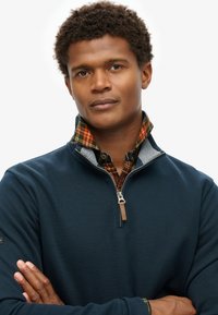 Navy-Fleecejacke mit Reißverschluss und karierter Kragen. Verfügt über strukturierten Stoff und einen silbernen Reißverschluss mit Lederzieher. Das Modell posiert mit verschränkten Armen.
