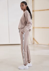 Beige Sweatshirt und passende Jogginghosen mit seitlichem Logo-Text; der Sweatshirt hat einen lockeren Schnitt, die Jogginghosen verfügen über einen elastischen Bund und Bündchen. Weiße Sneakers.