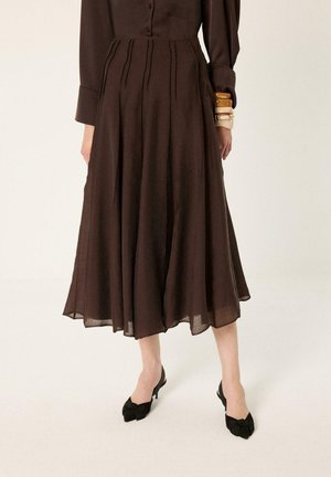 Brązowa plisowana spódnica midi w połączeniu z czarnymi czółenkami typu slingback na niskim obcasie oraz warstwową biżuterią na nadgarstku, noszona z dopasowanym brązowym topem.