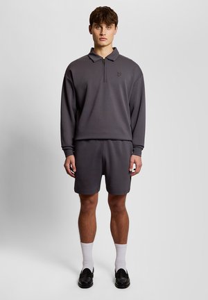 Young man stands facing forward wearing dark gray long-sleeve pullover with zipper, matching shorts, white socks, and black loafers on plain background.