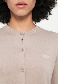 Bēša kardigans ar ribu apkakli, kas aprīkots ar divām pogām un nelielu "2ND" logotipu gaišā šuvumā. Mīksts adījuma tekstūra, cieši piegulošs dizains.