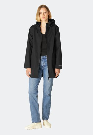 Zwarte hoodie met ritssluiting, lange mouwen en een strakke uitstraling; gedragen over een zwart topje en lichtblauwe jeans, gecombineerd met witte schoenen.