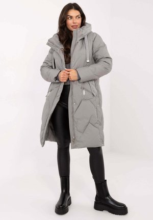 Long gray puffer coat with diagonal stitching, two front zippers, adjustable hood, and drawstring. Paired with black leggings and ankle boots.