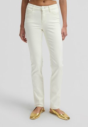 Kvinde iført højtaljede, hvide jeans med lige ben og metalliske guld flade sko med firkantet tå, stående foran en ensfarvet baggrund.