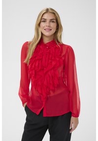 Femme portant un chemisier transparent rouge vif avec des volants superposés à l'avant, associé à un pantalon noir ample, souriant devant un fond uni.