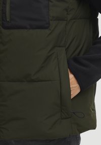 Dunkelgrüne gepolsterte Weste mit einer schwarzen Brusttasche, Reißverschluss, Seitentasche und verstellbarem Kordelzug am Saum. Weiches Fleecefutter ist sichtbar.