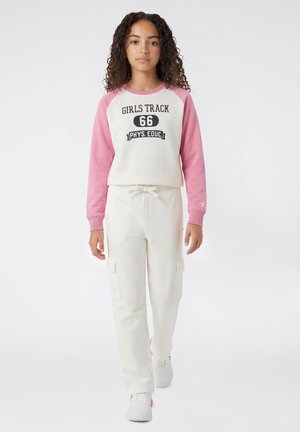 Mädchen mit lockigem Haar, das einen rosa-weißen "Girls Track 66 Phys. Educ." Pullover, weiße Cargo-Hosen und weiße Turnschuhe trägt und nach vorne geht.