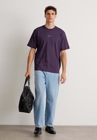 T-shirt en coton violet foncé à manches courtes avec un petit logo, associé à un jean bleu clair et des chaussures noires. Sac noir porté à la main.