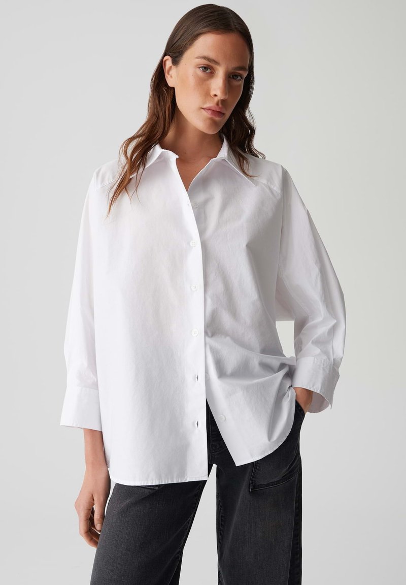 Vrouw met lang bruin haar, gekleed in een oversized witte blouse en zwarte broek, staand met één hand in de zak tegen een grijze achtergrond.